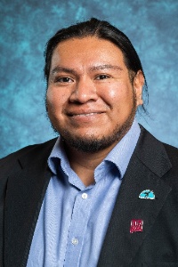 Picture of Michael Ray Director of American Indian Program. The image depicts a person with long, dark hair pulled back, dressed in formal attire. The individual is wearing a dark suit jacket over a light blue button-up shirt. There are two pins on the lapel of the suit jacket; one appears to have a teal design, and the other is a maroon rectangular pin with unidentified white text. The person has a trimmed beard and is smiling slightly. The background is a mottled blue and teal texture, giving a slightly abstract feel to the image.